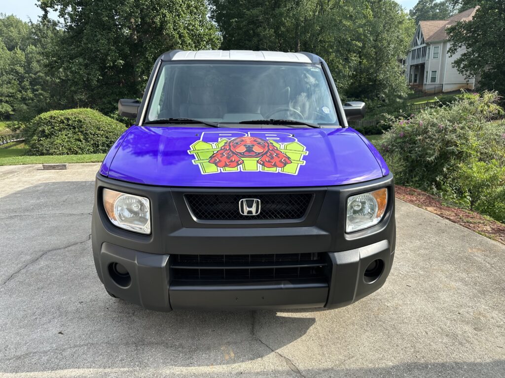 The Praline's Backyard dog taxi, a purple Honda Element with the company logo on the hood, parked in an Atlanta driveway