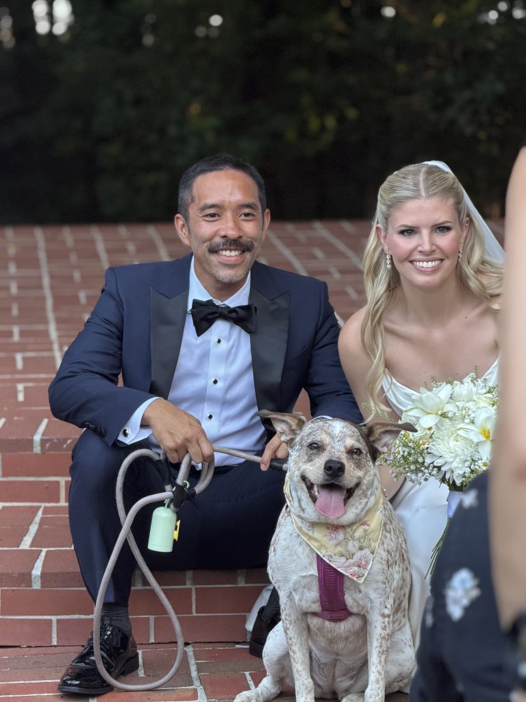 A bride and groom kneel on the venue steps beside their dog, who sits between them in a bandana.